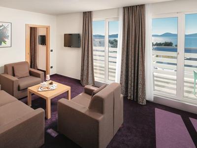 Lichte hotelkamer met bruine banken, houten tafel en groot raam met uitzicht op meer en bergen.