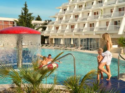 Hotelzwembad met waterspel, gasten in het water en vrouw met baby bij de rand.