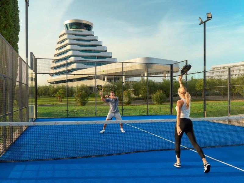 Twee vrouwen spelen tennis op een blauw tennisveld naast een modern hotelgebouw.