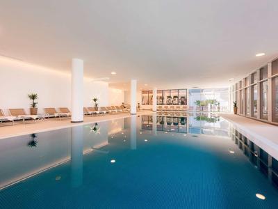 Großes Hallenbad mit blauem Wasser, weißen Säulen, Sitzbänken und großem Fensterfrontbereich.