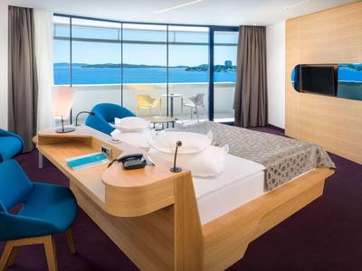 Modernes Hotelzimmer mit großem Bett, blauem Sessel und Panoramafenster mit Meerblick.