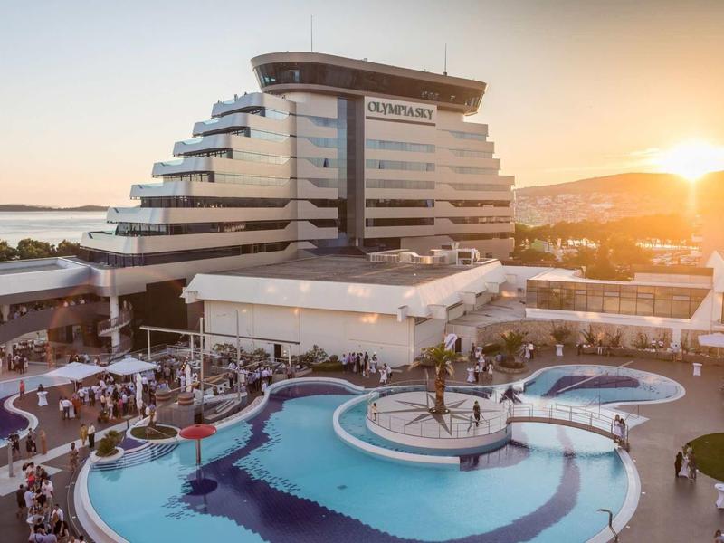 Modernes Hotel mit geschwungenem Pool und Sonnenschirmterrassen bei Sonnenuntergang am Wasser.