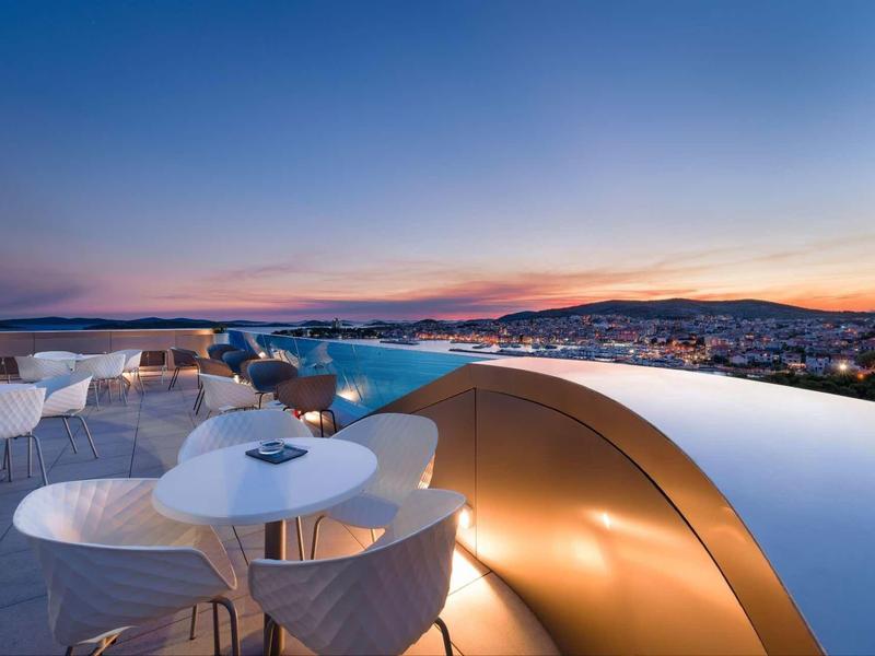 Moderne Rooftop-Bar mit runden weißen Tischen und Sesseln, Infinity-Pool, Blick auf Stadt bei Sonnenuntergang.