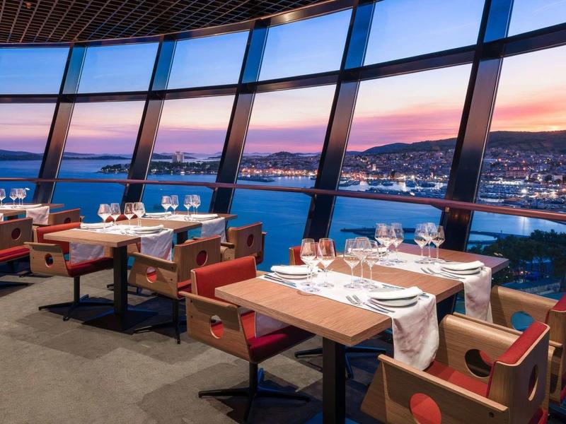 Modernes Restaurant mit großen Panoramafenstern, Holz- und Lederstühlen, Blick auf Stadt und Meer.