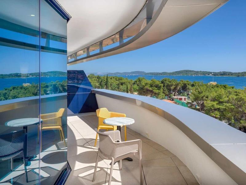 Moderner Balkon mit Glaswänden, zwei Stühlen und kleinem Tisch, Blick auf Meer und grüne Landschaft.