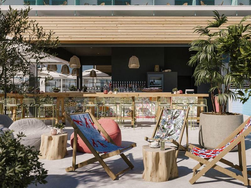 Außenbereich eines Cafés mit Holzliegen, Kissen, Stühlen und Pflanzen in warmem Sonnenlicht.