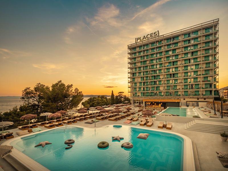 Moderner Hotelkomplex mit großem Outdoor-Pool, Sonnenliegen und Blick auf das Meer bei Sonnenuntergang.