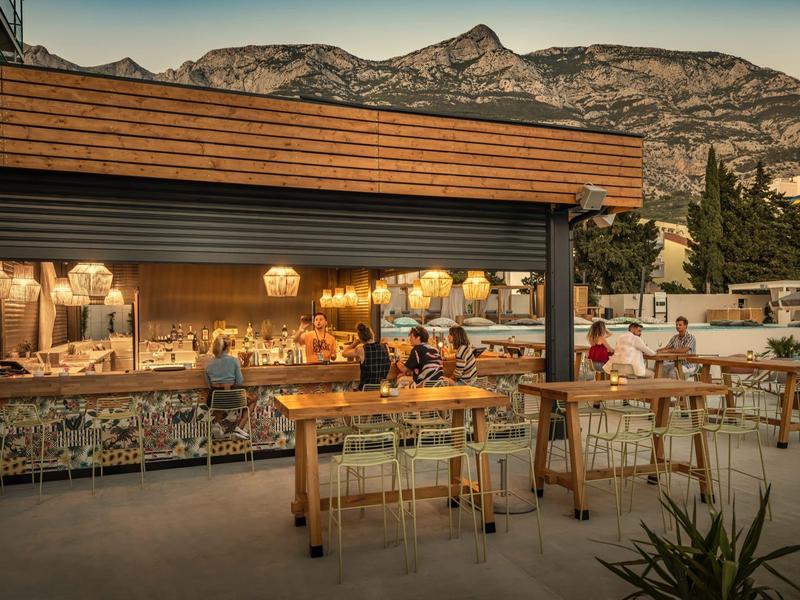 Outdoor-Bar mit Holztheke und Stehtischen vor Bergen und Abendhimmel bei Dämmerung.