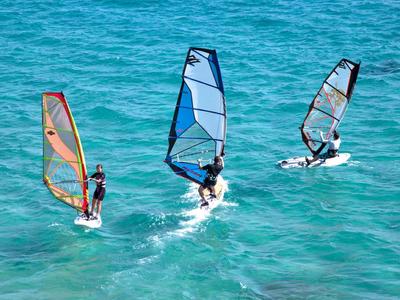Three windsurfers glide across the clear blue sea water on a sunny day.