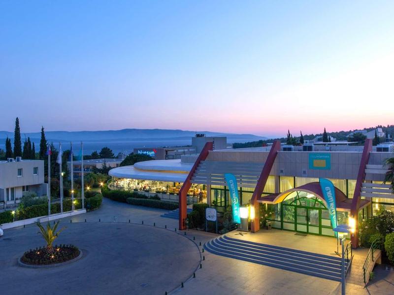 Moderní hotelová budova při západu slunce s osvětlenou terasou a výhledem na moře.