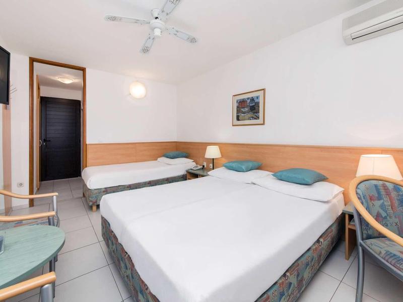 Hotelový pokoj s manželskou postelí, jednolůžkem, dvěma židlemi a televizí na zdi.