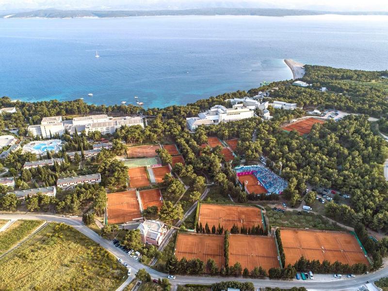 Letecký pohled na hotel s tenisovými kurty obklopený lesem a mořem v pozadí.