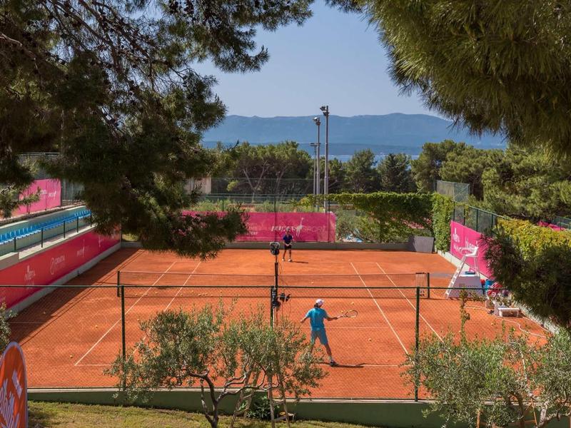 Tenisový kurt s červenou hlínou, hráči, výhledem na moře a okolními stromy.