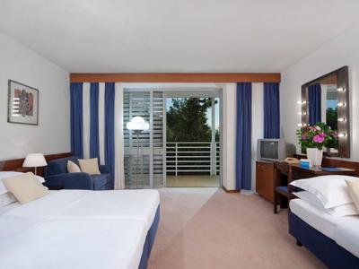 Světý hotelový pokoj se dvěma postelemi, pohovkou, psacím stolem a balkónem s výhledem do zeleně.