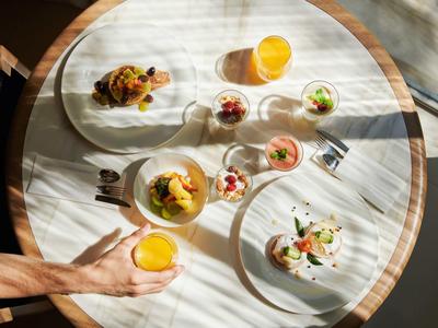 Ein runder Holztisch mit gedecktem Frühstück und zwei Gläsern Orangensaft, eine Hand nimmt ein Glas.