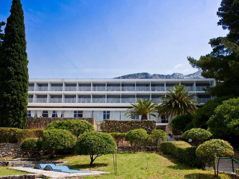 Moderní vícepodlažní hotel s balkony, obklopený zahradou s stromy a keři.