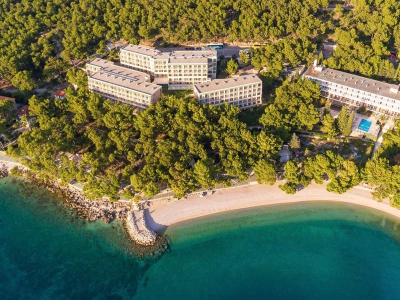 Hotelový areál obklopený hustým lesem u zakřivené písečné zátoky s čistou modrou vodou.