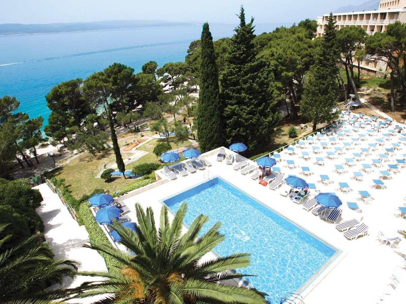 Hotelový bazén s modrými slunečníky a lehátky obklopený stromy u moře.