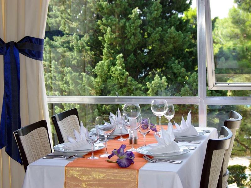 Stůl prostřený s bílými ubrusy a sklenicemi u okna s výhledem na zelené stromy.