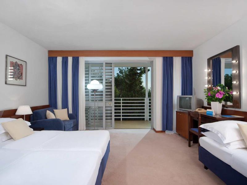 Světý hotelový pokoj se dvěma postelemi, pohovkou, psacím stolem a balkónem s výhledem do zeleně.