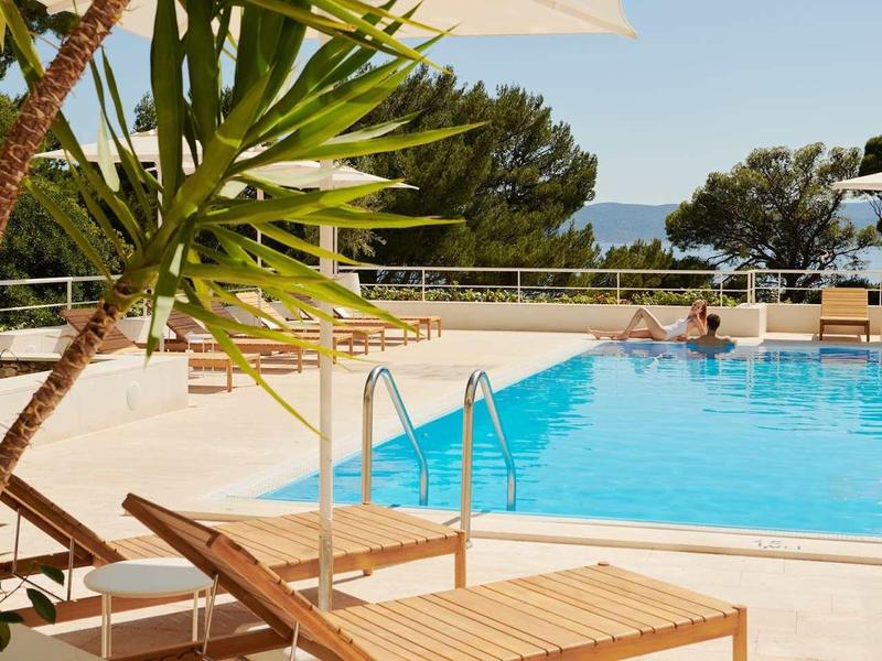 Velký venkovní bazén s lehátky a výhledem na moře v pozadí.