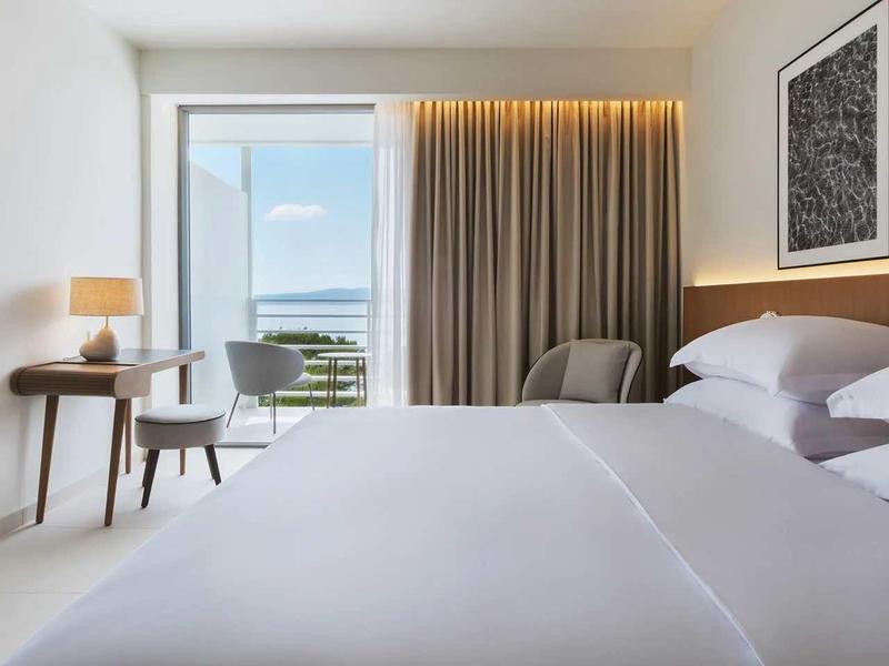 Moderní hotelový pokoj s velkou postelí, pracovním stolem a výhledem na moře oknem.