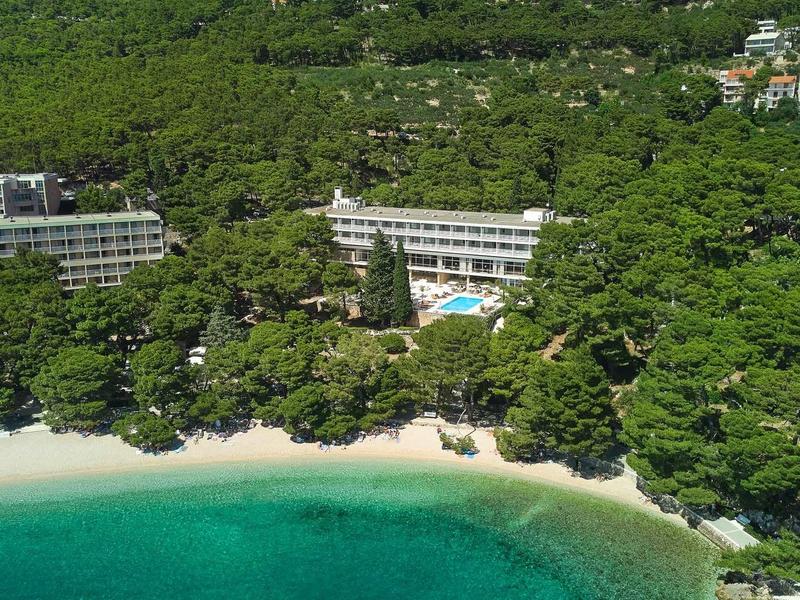 Hotelový komplex s bazénem na pláži, obklopený hustou zelení a tyrkysovou vodou.