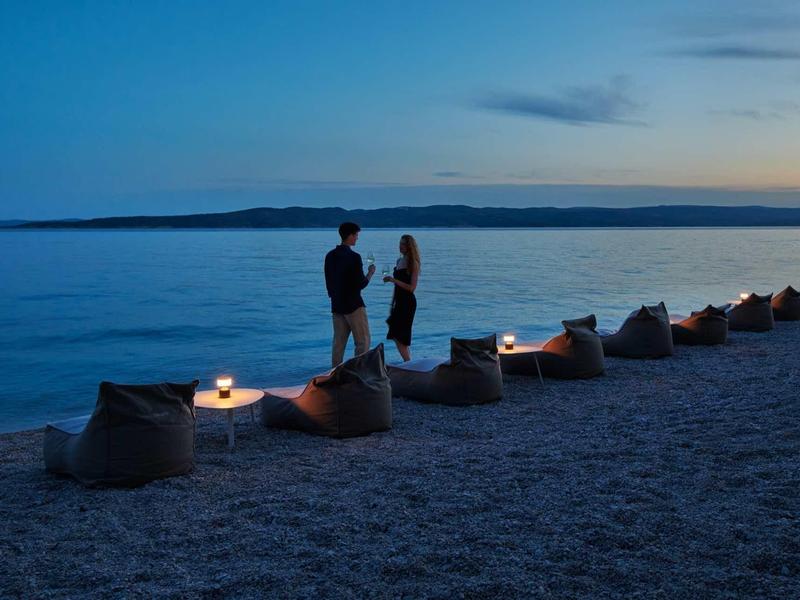 Dva lidé stojí na pláži u světla svíček a pozorují klidný oceán za soumraku.