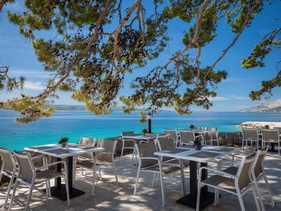 Restaurante al aire libre con mesas y sillas blancas bajo ramas de árboles con vista al mar azul.