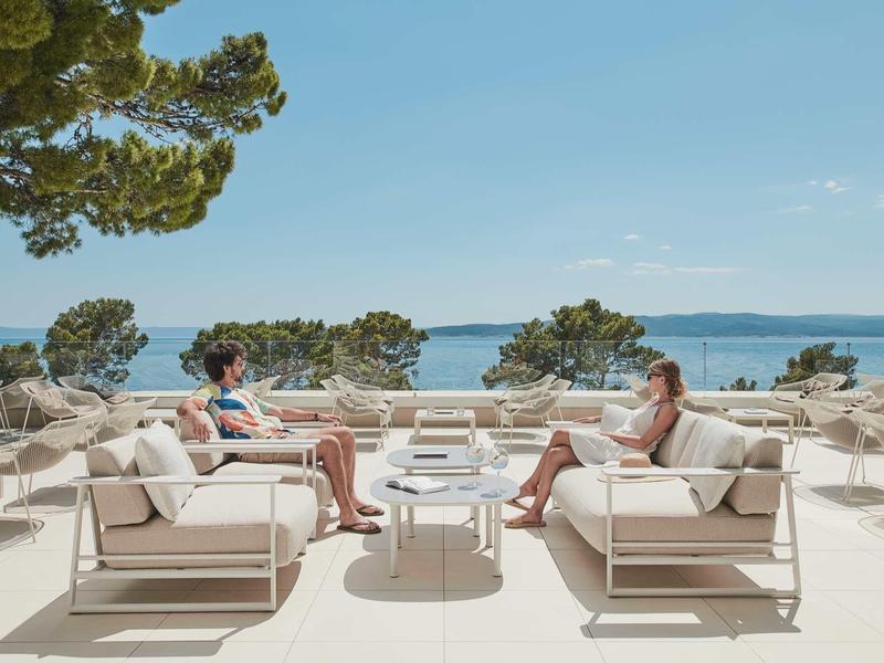 Deux personnes se détendent sur des canapés blancs avec vue sur la mer sur une terrasse d'hôtel.