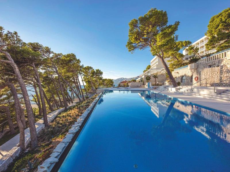 Piscine à débordement avec vue sur la mer, entourée d'arbres et de rochers sous un ciel clair.