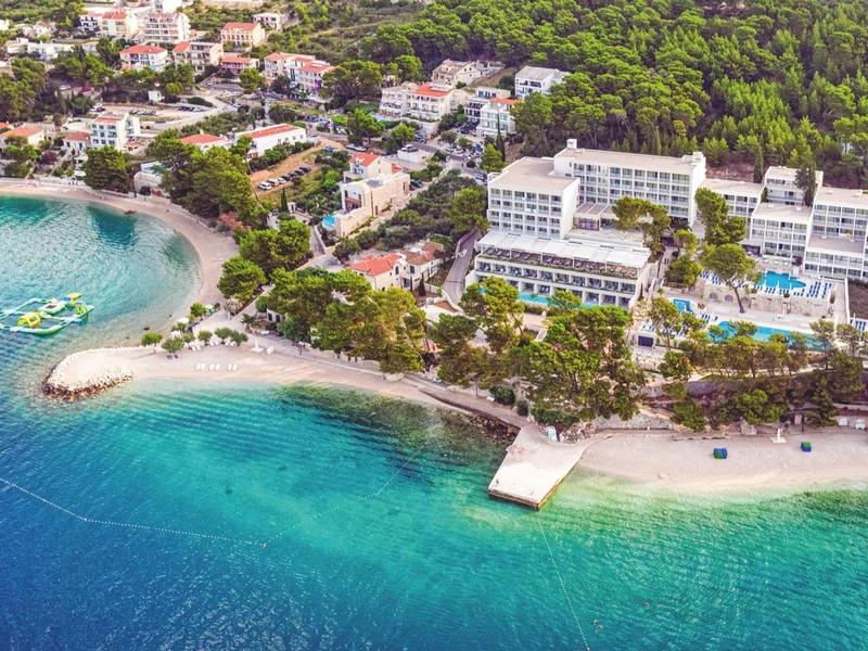 Hôtel de luxe avec piscine au bord de la mer turquoise, entouré de forêt verte et plage de sable.