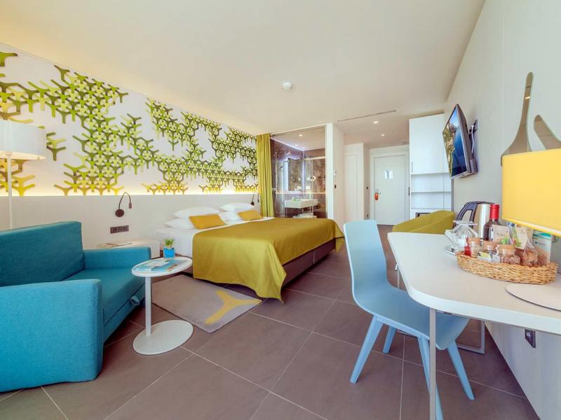 Chambre d'hôtel moderne avec lit double, fauteuil bleu, bureau et décoration murale jaune.