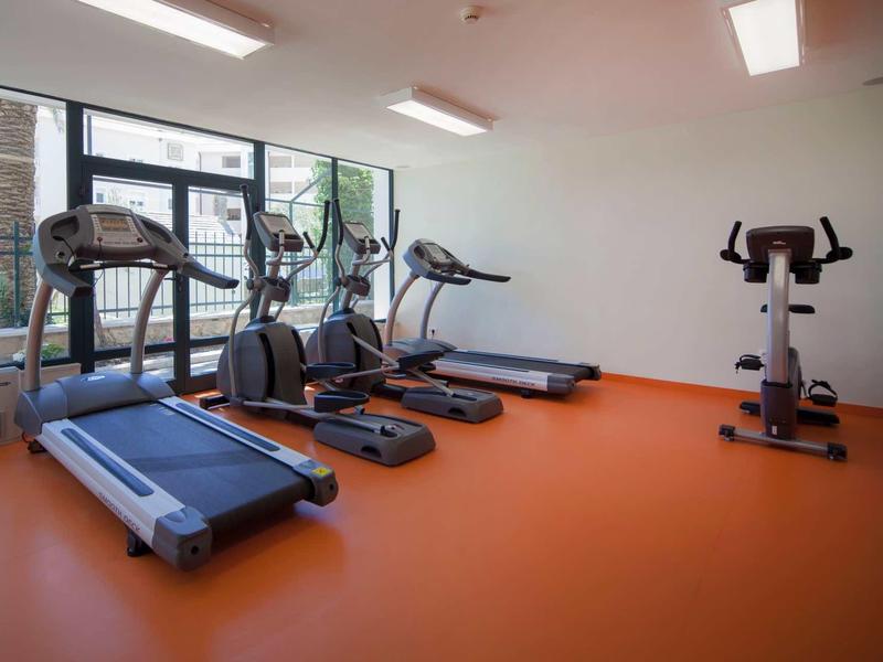 Jasno oświetlona siłownia z bieżniami, orbitrekami i rowerem na pomarańczowej podłodze.