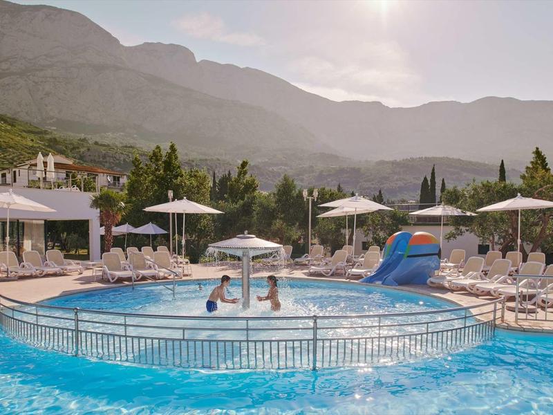 Duży basen z parasolami i leżakami na tle górskiego krajobrazu o zachodzie słońca.