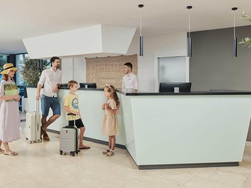 Rodzina z dziećmi i bagażem przy recepcji hotelu gotowa do zameldowania.