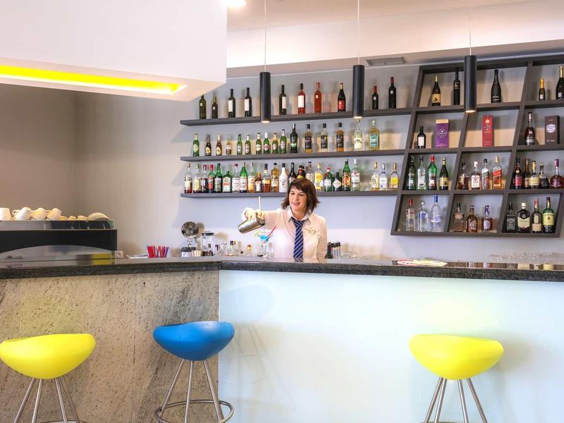 Nowoczesny bar hotelowy z kolorowymi stołkami i barmanem przygotowującym napoje.