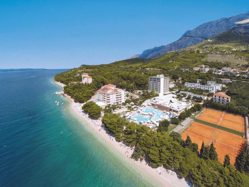 Widok z lotu ptaka na nadmorski kurort z plażą, budynkami hotelowymi, basenami i kortami tenisowymi przy morzu.