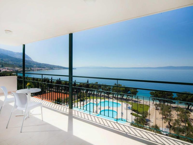 Balkon z białymi krzesłami z widokiem na basen i morze pod niebieskim niebem.