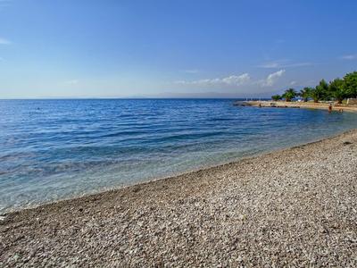 Kiesstrand mit ruhigem blauem Meer und klarem Himmel, bewaldeter Bereich rechts im Hintergrund.