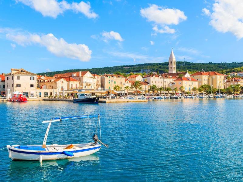 Kleines Boot auf stillem blauem Wasser vor bunten Häusern und Hügeln bei sonnigem Himmel.