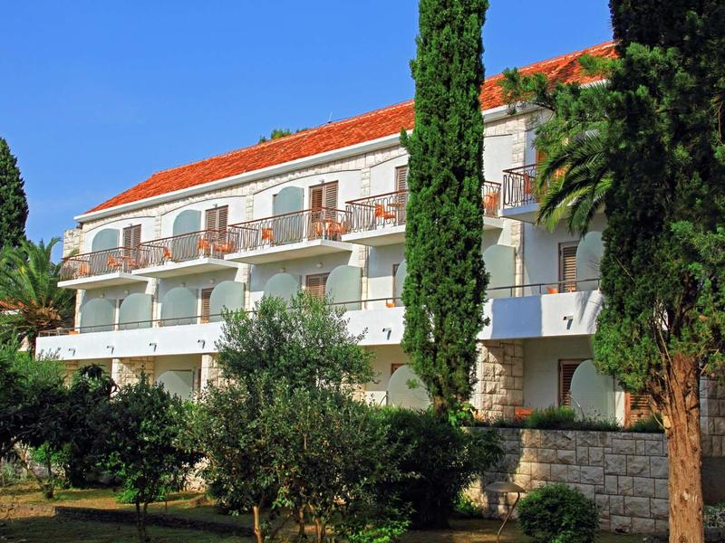 Mehrstöckiges Haus mit roten Dachziegeln, weißen Balkonen und hohen grünen Bäumen im Vorgarten.