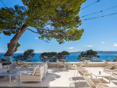 Terrassenbereich mit weißen Sitzmöbeln, großen Bäumen und Blick auf das blaue Meer unter klarem Himmel.