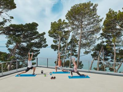 Drei Menschen trainieren auf blauen Matten im Freien mit Blick auf das Meer und Bäume.