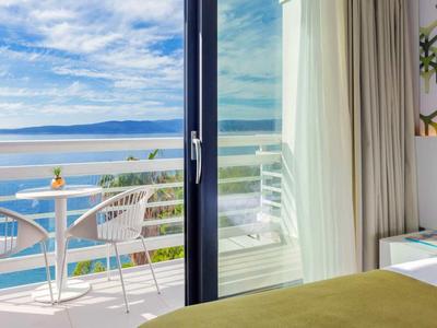 Helle Hotelzimmertür öffnet sich zu einem Balkon mit Meerblick und weißem Mobiliar.