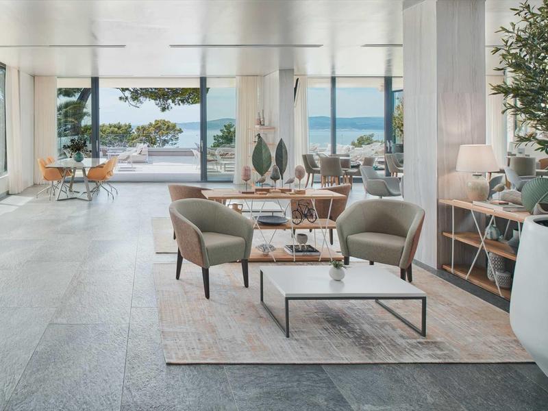 Lichte hotellounge met fauteuils, tafels en uitzicht door grote ramen op terras en zee.