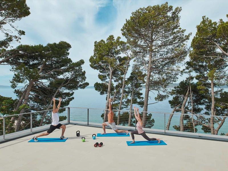 Drie vrouwen doen yoga op matten op een terras met uitzicht op bomen en de zee.