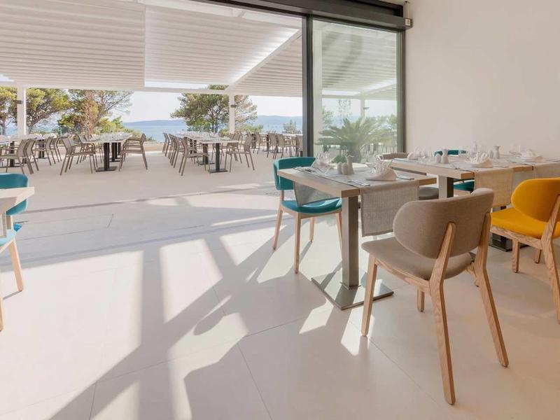 Licht modern restaurant met kleurrijke stoelen en uitzicht op een zonnig terras.