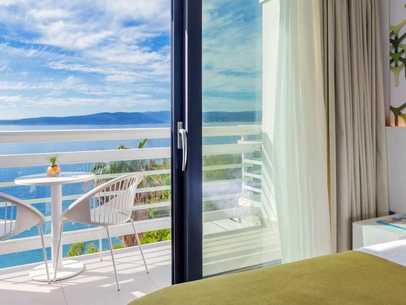 Lichte hotelkamer met balkon, zeezicht en moderne meubels bij daglicht.