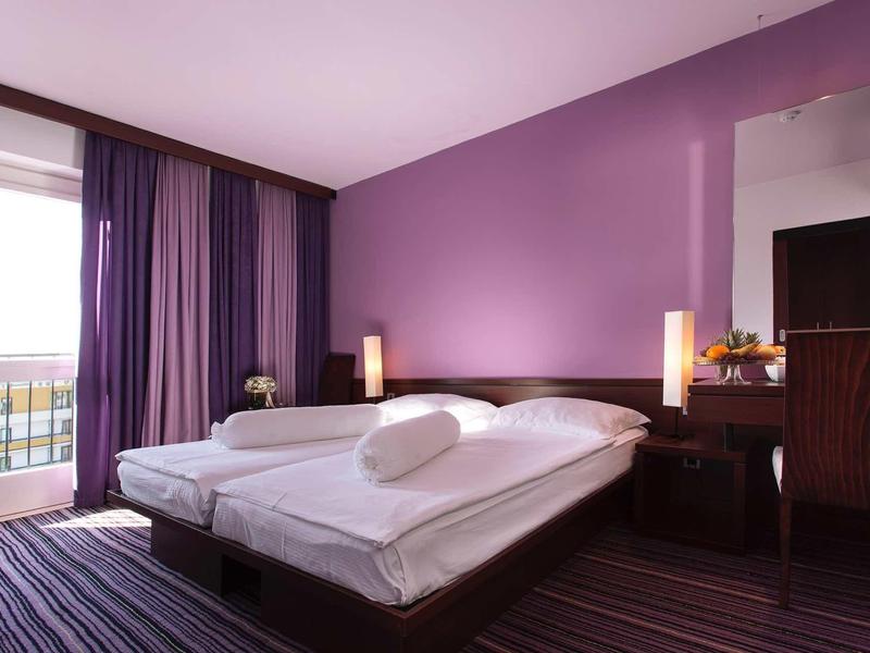 Hotelový pokoj s fialovými stěnami, manželskou postelí, psacím stolem a balkonem s výhledem ven.